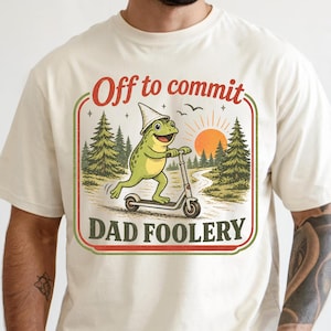 May include: Cream-coloured t-shirt with a cartoon frog on a scooter. The frog wears a hat, set against trees and a rising sun. Text reads "Off to commit DAD FOOLERY".