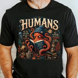 May include: A black t-shirt featuring a graphic of an orange octopus wearing a blue knit cap and reading a book titled 'Remarkably Bright Creatures'. The design includes text reading 'HUMANS', 'Good Day Sir', and 'Day 1,299' with underwater elements.