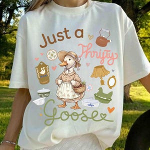 May include: Cream-colored t-shirt with the text "Just a Thrifty Goose" and a cartoon goose wearing a bonnet and apron. The shirt features various illustrations of kitchenware and hearts, creating a whimsical, vintage aesthetic.