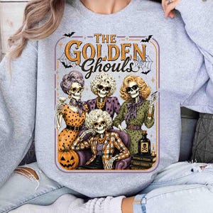 May include: A light grey crewneck sweatshirt featuring a graphic print of four skeletons styled as older women. The text reads THE GOLDEN Ghouls. The skeletons are dressed in patterned clothing, sitting with a pumpkin, a bottle, and books.