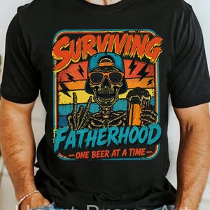 Könnte beinhalten: Schwarzes T-Shirt mit einer farbenfrohen Grafik, die ein Skelett mit Sonnenbrille und Baseballkappe zeigt, das ein Bier hält. Der Text lautet "SURVIVING FATHERHOOD - ONE BEER AT A TIME."