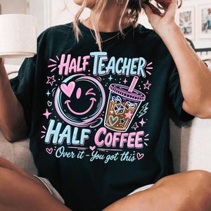 May include: A black t-shirt featuring a graphic design with the text "Half Teacher Half Coffee" and "Over it - You got this". The design includes a winking smiley face with a pink heart eye and an iced coffee cup with ice cubes and stars.