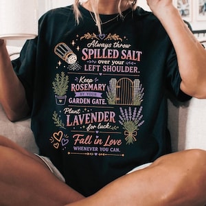 以下が含まれることがあります： 黒の半袖Tシャツ。グラフィックには「Always throw SPILLED SALT OVER YOUR LEFT SHOULDER. Keep ROSEMARY BY YOUR GARDEN GATE. Plant LAVENDER for luck. Fall in Love WHENEVER YOU CAN.」という文字と、塩、ローズマリー、門、ラベンダーのイラストが描かれています。