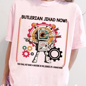Puede incluir: Camiseta rosa claro con una ilustración de robot de estilo retro y el texto "BUTLERIAN JIHAD NOW!" y "THOU SHALL NOT MAKE A MACHINE IN THE LIKENESS OF A HUMAN MIND". El diseño incluye engranajes, flechas y bombillas.