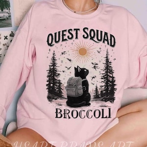 May include: A light pink sweatshirt with the words "QUEST SQUAD BROCCOLI" printed on the front. The graphic features a black cat wearing a backpack, set against a forest scene with a sun and flying creatures.