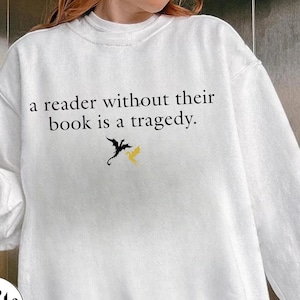 Op de afbeelding: Witte trui met de tekst "a reader without their book is a tragedy." en een zwarte en gele drakenafbeelding.