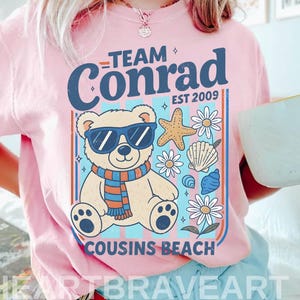 May include: Pink t-shirt with a graphic featuring a bear wearing sunglasses and a scarf, surrounded by seashells and flowers. Text reads "TEAM Conrad EST 2009" and "COUSINS BEACH". The shirt is from HEARTBRAVEART.