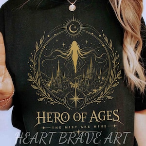May include: Black t-shirt with a gold graphic design featuring a silhouette of a figure, a crescent moon, a cityscape, and the text "HERO OF AGES". The text "THE MIST ARE MINE" and "HEART BRAVE ART" are also included.