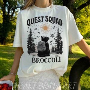May include: Off-white t-shirt with a graphic featuring a black cat wearing a backpack, set against a forest scene with the text "QUEST SQUAD BROCCOLI". The design includes a sun, birds, and trees.
