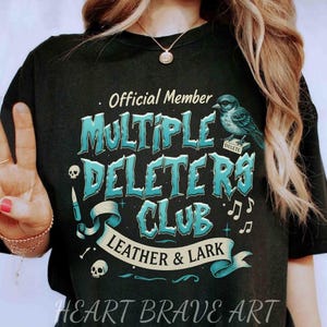 May include: Black t-shirt with a blue and white graphic design that says "Official Member Multiple Deleters Club Leather & Lark" with a bird and musical notes. The text "Heart Brave Art" is at the bottom of the shirt.