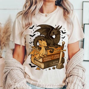 May include: Beige t-shirt featuring a dragon graphic. Two dragons are perched on a stack of books with titles like "Rebecca Yarros" and "Iron Flame." A sword and bats complete the fantasy design.