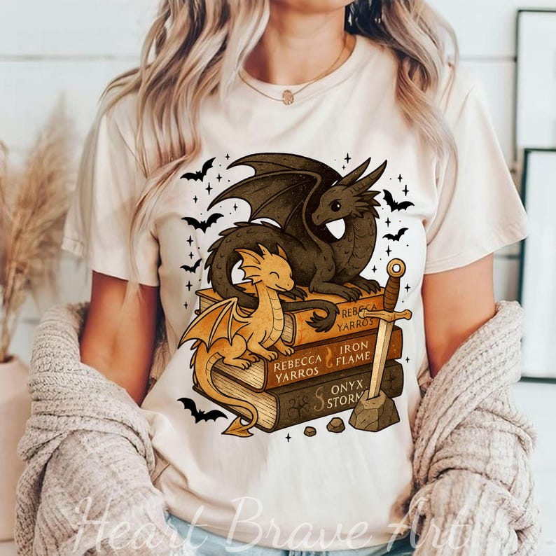 May include: Beige t-shirt featuring a dragon graphic. Two dragons are perched on a stack of books with titles like "Rebecca Yarros" and "Iron Flame." A sword and bats complete the fantasy design.