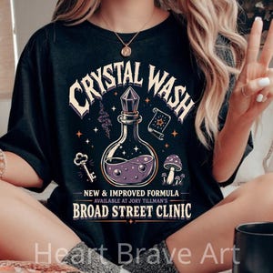 May include: A black t-shirt featuring a graphic print of a potion bottle filled with purple liquid, a key, a scroll, and mushrooms. The text reads CRYSTAL WASH, NEW & IMPROVED FORMULA, AVAILABLE AT JORY TILLMAN'S BROAD STREET CLINIC.