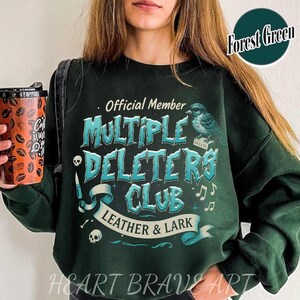 May include: A dark green sweatshirt with the text "Official Member Multiple Deletors Club Leather & Lark" in a blue and white font. The text is surrounded by a decorative banner and a small bird graphic. The text "Forest Green" is in a white box in the upper right corner of the image.
