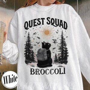 May include: White sweatshirt with a graphic featuring a black cat wearing a backpack, set against a forest scene with a sun and flying creatures. The text "QUEST SQUAD BROCCOLI" is printed above and below the cat.