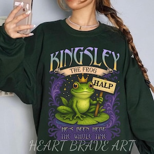 Puede incluir: Sudadera verde oscuro con un dibujo de una rana de dibujos animados con una corona y un cartel que dice "HALP". La sudadera tiene el texto "KINGSLEY THE FROG" y "HE'S BEEN HERE THE WHOLE TIME" con "HEART BRAVE ART" en la parte inferior.