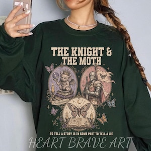 May include: A dark green long-sleeve sweatshirt featuring a graphic print with the text THE KNIGHT & THE MOTH and TO TELL A STORY IS IN SOME PART TO TELL A LIE. The design includes a gargoyle, a knight in armour, and a moth over an open book.
