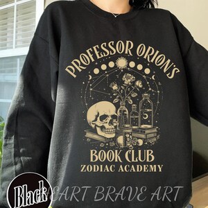 May include: Black sweatshirt with a graphic design featuring a skull, flowers, and bottles. The text reads "Professor Orion's Book Club Zodiac Academy".