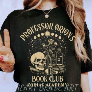 Puede incluir: Camiseta negra con un diseño gráfico dorado que presenta una calavera, flores, botellas y libros. El texto "Professor Orion's Book Club Zodiac Academy" está impreso en la camiseta.
