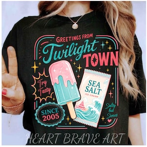 Op de afbeelding: Een zwart t-shirt met een kleurrijke afbeelding met de tekst Greetings From Twilight Town. Het ontwerp bevat een roze en blauw ijsje, een verpakking van zeezoutijs met oceaangolven en de tekst Try Tasty, Since 2005, en Salty but Sweet.