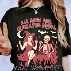 Puede incluir: Camiseta negra con un diseño de Halloween. El diseño presenta una bruja, un esqueleto, murciélagos y el texto "ALL MEN ARE CREMATED EQUAL". La camiseta también tiene el texto "HEART BRAVE ART" en la parte inferior.