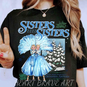 May include: Black t-shirt with a graphic of two women in blue dresses holding a blue fan, with the word "Sisters" in a decorative font. The image includes a winter scene and the words "Heart Brave Art".