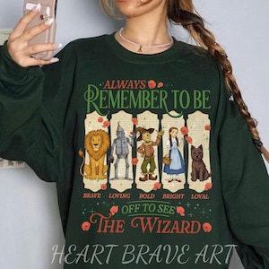 Op de afbeelding: Donkergroene sweatshirt met een afbeelding van personages uit The Wizard of Oz, waaronder een leeuw, een tinnen man, een vogelverschrikker, Dorothy en Toto. Tekst luidt "ALWAYS REMEMBER TO BE" en "OFF TO SEE THE WIZARD".