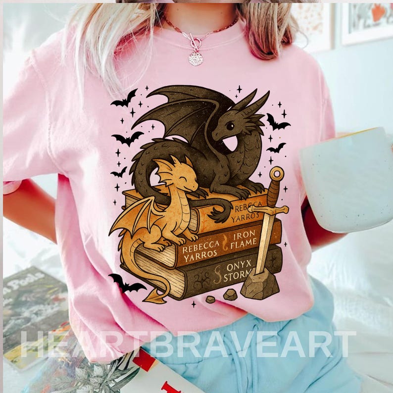 May include: Pink t-shirt featuring a graphic of two dragons atop a stack of books. The books are titled "Rebecca Yarros", "Iron Flame", and "Onyx Storm". A sword is placed next to the books, and bats and stars surround the dragons.