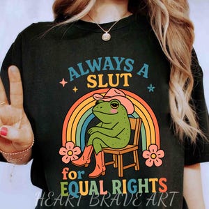 May include: Black t-shirt with a graphic of a frog wearing a cowboy hat and boots, sitting in a chair in front of a rainbow. Text reads "ALWAYS A SLUT for EQUAL RIGHTS".