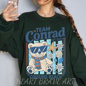 May include: Dark green sweatshirt with a graphic featuring a bear wearing sunglasses and a scarf, with the text "Team Conrad" and "Cousins Beach". The design includes a starfish, flowers, and seashells. The text "Heart Brave Art" is at the bottom.