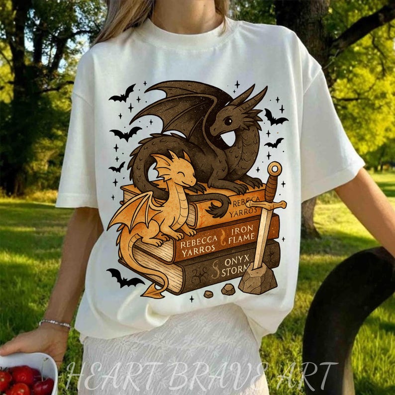May include: White t-shirt featuring a dragon design. The graphic includes two dragons, one brown and one orange, perched on a stack of books. The books are titled "Rebecca Yarros", "Iron Flame", and "Onyx Storm". Bats and stars surround the dragons.