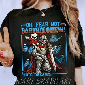 May include: Black t-shirt with a graphic featuring a knight in shining armor, a small vampire, and blue butterflies. Text reads "OH, FEAR NOT BARTHOLOMEW! HE'S DREAMY." The design is framed in a blue border. "HEART BRAVE ART" is printed at the bottom.