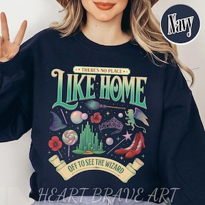 以下が含まれることがあります： 「THERE'S NO PLACE LIKE HOME」と「OFF TO SEE THE WIZARD」の文字が入った、カラフルなグラフィックデザインのネイビーブルーのスウェットシャツ。デザインには、魔女の帽子、赤い靴、ロリポップのイラストが含まれています。「HEART BRAVE ART」のブランド名が下部にあります。