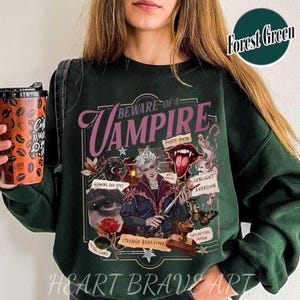 Op de afbeelding: Een donkergroen sweatshirt met een grafische print van een vampier. De grafiek bevat de tekst "Beware of a Vampire" en toont verschillende kenmerken van vampiers, zoals puntige tanden, gloeiende rode ogen, afkeer van zonlicht en vreemd gedrag.