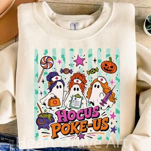 May include: A cream-colored crewneck sweatshirt featuring a graphic of three ghosts dressed as nurses. The design includes a lollipop, a pumpkin, a purple cauldron, and the text HOCUS POKE-US in bold orange and purple letters on a striped background.