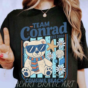 May include: Black t-shirt with a graphic featuring a bear wearing sunglasses and a scarf, surrounded by beach-themed elements. The text "Team Conrad EST 2009" and "Cousins Beach" are also displayed.