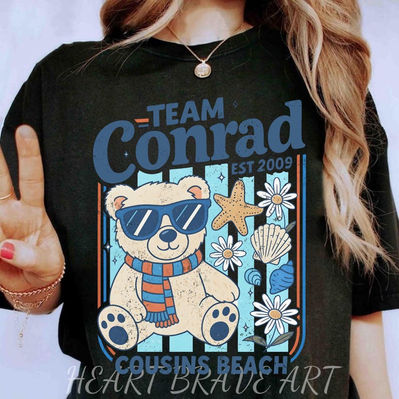 May include: Black t-shirt with a graphic featuring a bear wearing sunglasses and a scarf, surrounded by beach-themed elements. The text "Team Conrad EST 2009" and "Cousins Beach" are also displayed.