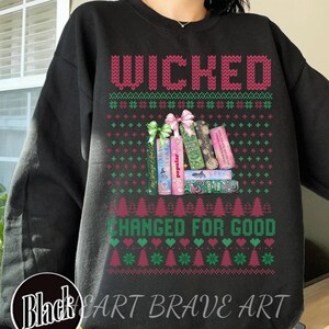 Könnte beinhalten: Schwarzes Sweatshirt mit den Worten "WICKED" und "CHANGED FOR GOOD" in einem festlichen Design im Sweater-Stil. Das Design umfasst gestapelte Bücher mit Titeln wie "Popular" und "For Good". Das Sweatshirt hat auch ein Logo mit der Aufschrift "Black Cart Brave Art".