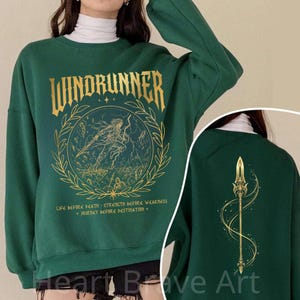 May include: A dark green sweatshirt featuring a gold "Windrunner" graphic with a person and bow inside a wreath. The back displays a gold spear design. The text below the front graphic reads "Life before death..." and "Journey before destination".