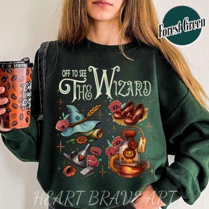 Könnte beinhalten: Dunkelgrünes Sweatshirt mit dem Text "Off to See The Wizard" und Illustrationen eines Hutes, roter Schuhe und eines Löwen. Das Design umfasst rote Mohnblumen und Sterne. Das Sweatshirt ist dunkelgrün.