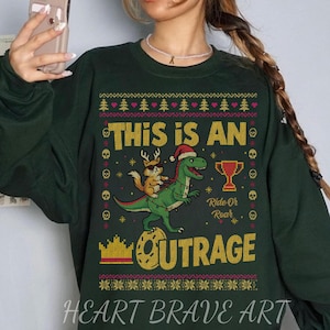 May include: Dark green sweatshirt with a festive graphic. The design features a green dinosaur wearing a Santa hat, ridden by a small brown animal. Text includes "THIS IS AN OUTRAGE" and "Ride Or Roar". The words "HEART BRAVE ART" are at the bottom.