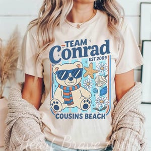 May include: Beige t-shirt featuring a graphic of a bear wearing sunglasses and a scarf, with the text "Team Conrad" and "Cousins Beach". The design includes starfish, seashells, and flowers, with the year "EST 2009".