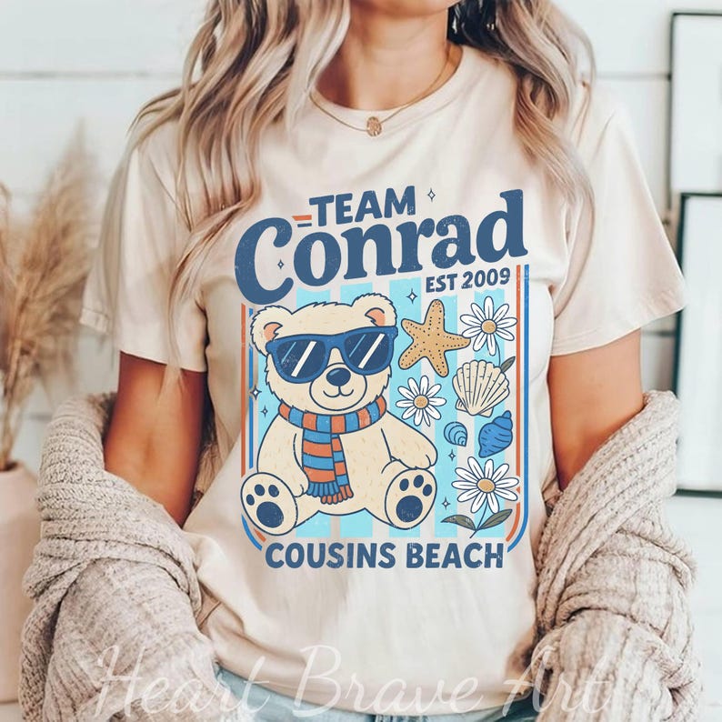 May include: Beige t-shirt featuring a graphic of a bear wearing sunglasses and a scarf, with the text "Team Conrad" and "Cousins Beach". The design includes starfish, seashells, and flowers, with the year "EST 2009".