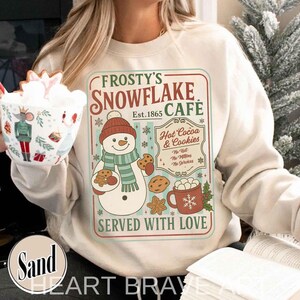 Puede incluir: Sudadera color crema con un diseño de Frosty's Snowflake Cafe. El gráfico incluye un muñeco de nieve, galletas y una taza de chocolate caliente. El texto dice "Served with Love."