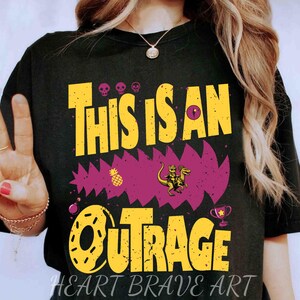 Puede incluir: Camiseta negra con el texto "THIS IS AN OUTRAGE" en amarillo, con un dise&ntilde;o p&uacute;rpura dentado. La camiseta presenta una pi&ntilde;a, un dinosaurio montando otro dinosaurio y un trofeo. Las palabras "HEART BRAVE ART" est&aacute;n en la parte inferior.