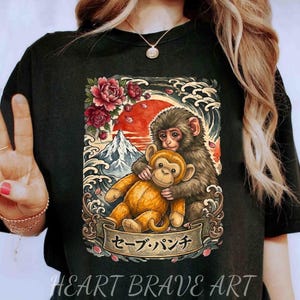 May include: Black t-shirt with a graphic of two monkeys, one embracing a plush toy monkey. The design incorporates a red sun, mountain, and floral details, with Japanese text on a banner. The text "HEART BRAVE ART" is at the bottom.