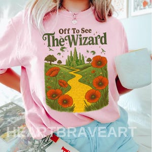 Könnte beinhalten: Hellrosa Rundhals-T-Shirt mit einem Aufdruck einer gelben Ziegelsteinstraße, die zu einer grünen Stadt führt, umgeben von roten Mohnblumen. Der Text "Off To See The Wizard" ist grün. Das Shirt hat einen Vintage-Look.