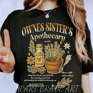 Puede incluir: Camiseta negra con un diseño gráfico que presenta el texto "OWNES SISTER'S Apothecary est. 1995". El diseño incluye ilustraciones de una botella, mortero y maja, hierbas y texto. Las palabras "HEART BRAVE ART" están en la parte inferior.