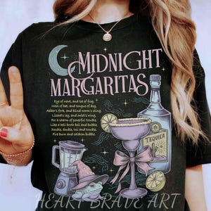 May include: Black t-shirt with a whimsical "Midnight Margaritas" design. The graphic includes a margarita glass, tequila bottle, blender, and a frog wearing a witch hat. The text includes a potion recipe.