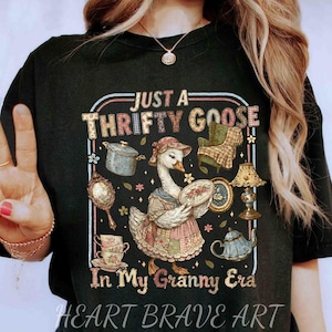 Puede incluir: Camiseta negra con un gráfico de un ganso con ropa vintage sosteniendo un plato, rodeado de objetos antiguos. El texto dice "Just a Thrifty Goose In My Granny Era" y "Heart Brave Art".